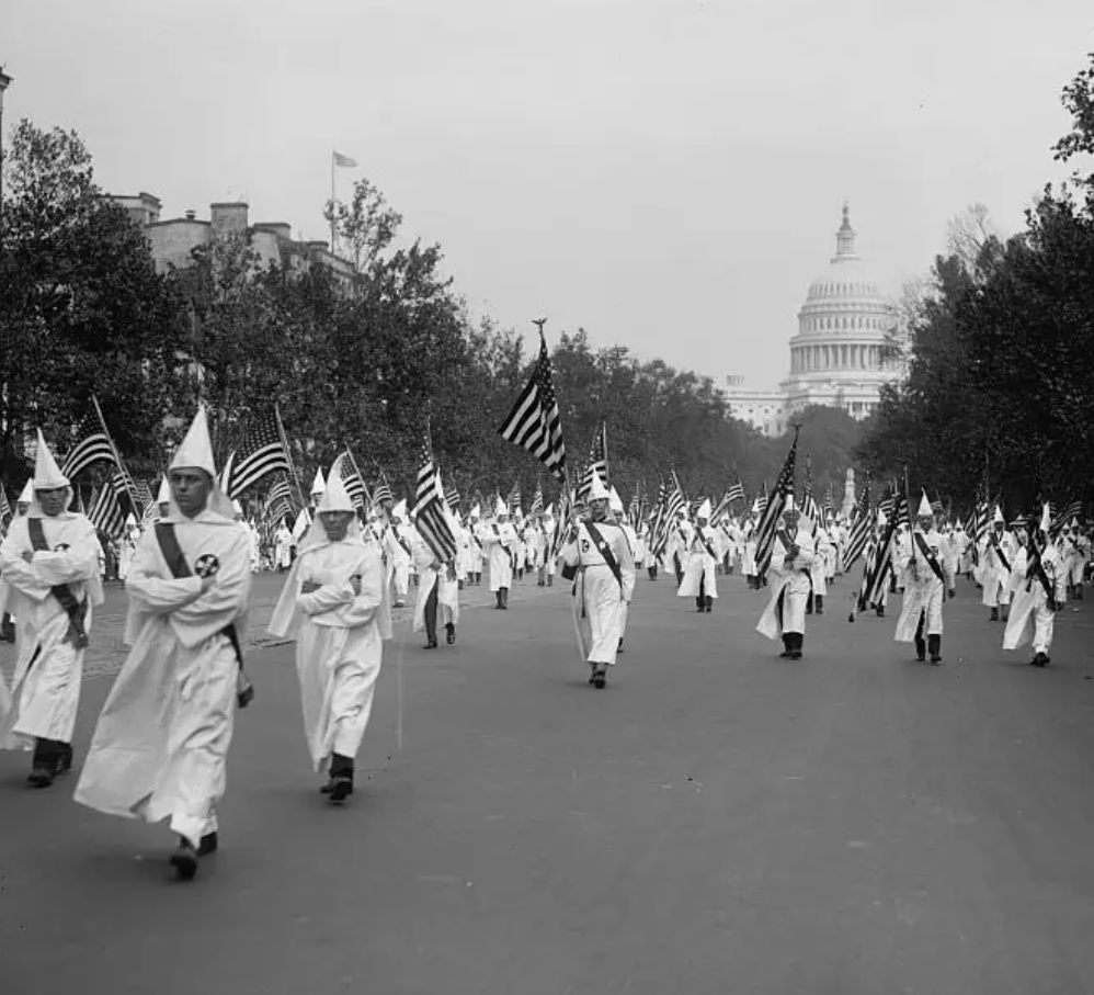 an archival black and white photograph of the KKK marching in Washington DC with the capitol behind them