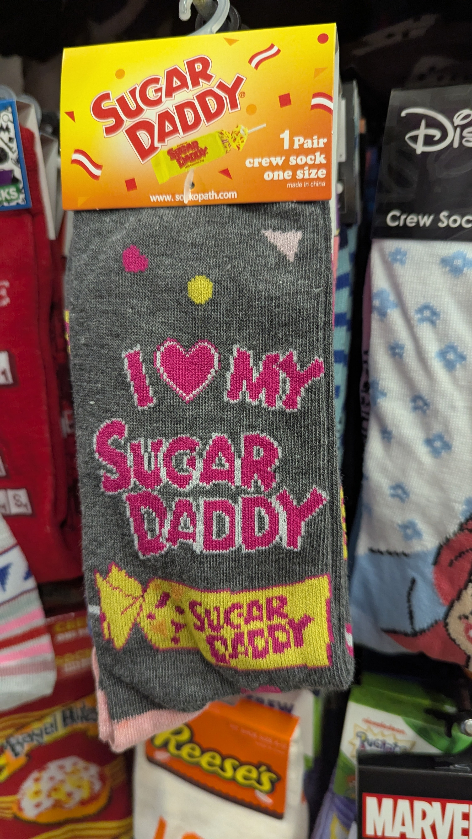 A pair of gray socks with colorful text and designs featuring the slogan I ♥ My Sugar Daddy along with candy graphics, displayed in a store.