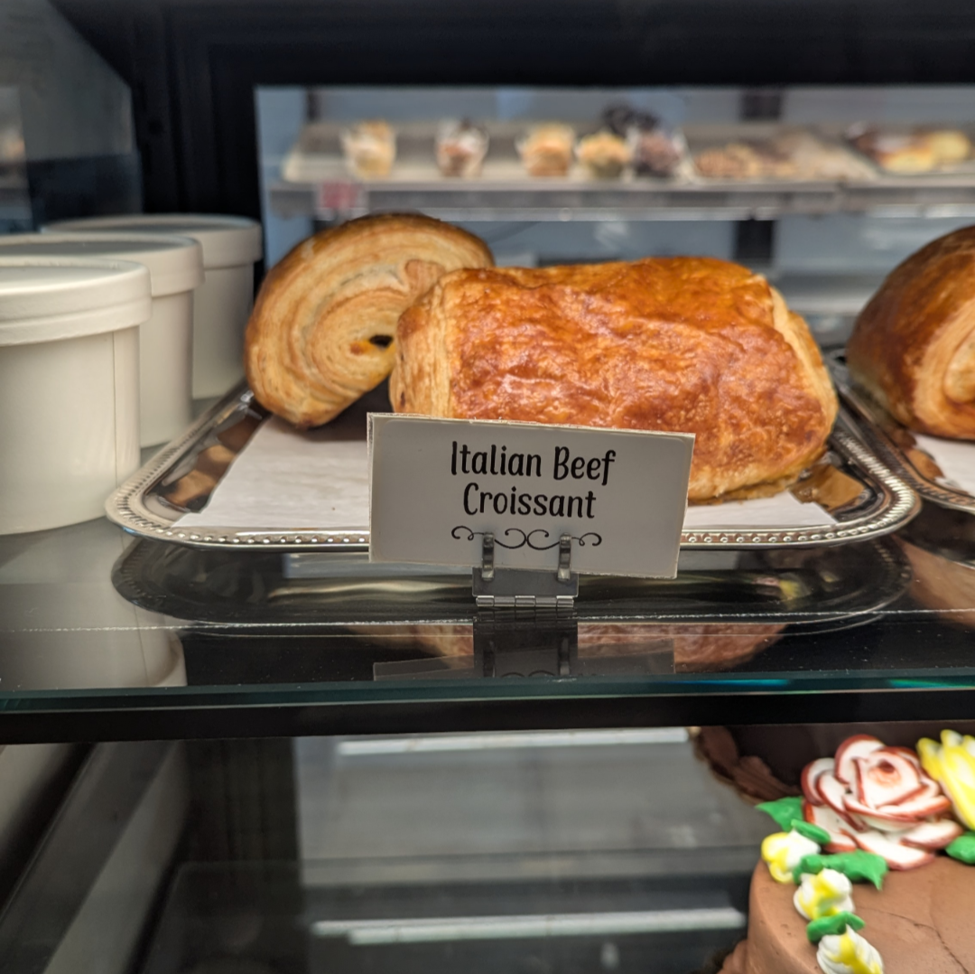 A display case contains Italian beef croissants, other pastries, and a decorated cake.