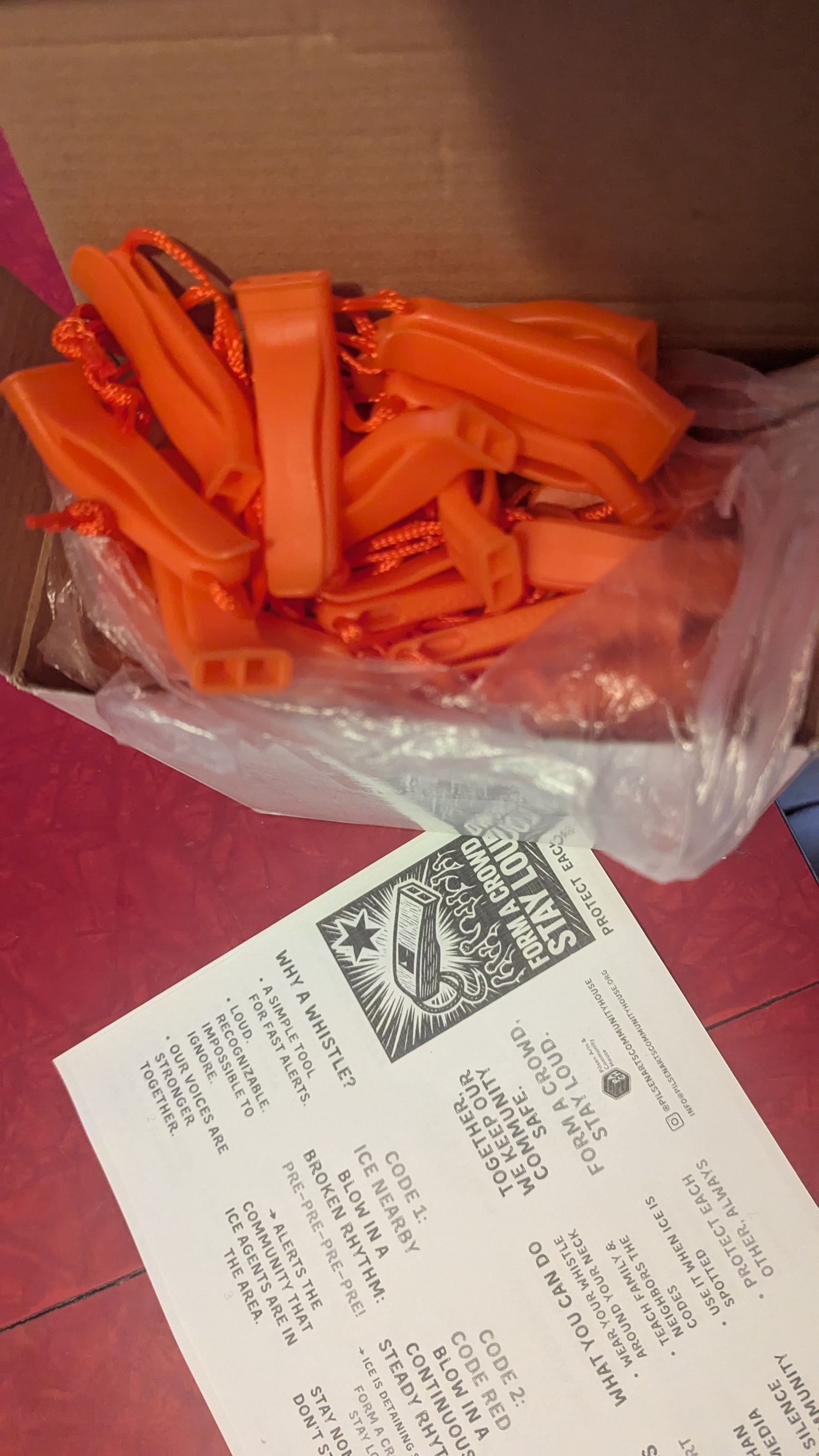 A box containing several orange whistles with cords next to a printed sheet of information on how to use them to warn about ICE