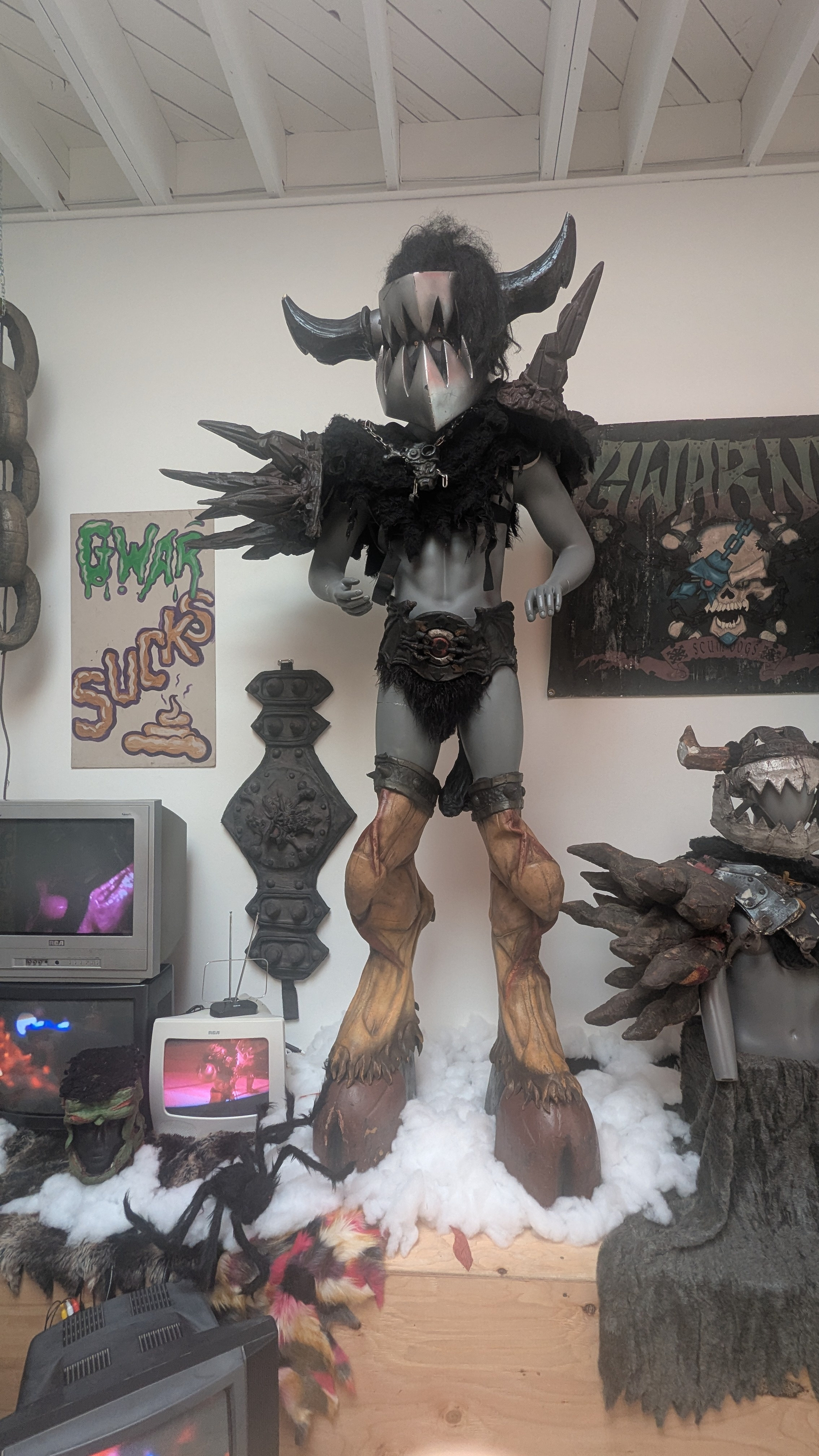 A room features a large fantasy monster statue with horns and hoof-like feet, surrounded by various artworks, televisions, and a sign that reads GWAR SUCKS.