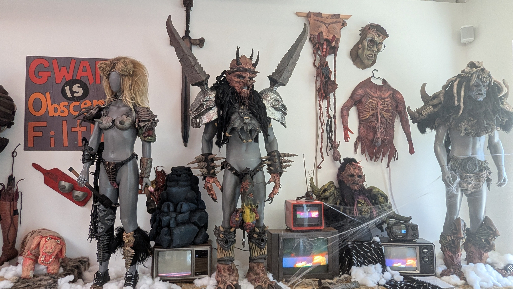 A display showcases mannequins dressed in elaborate, fantastical costumes alongside TV sets and various props, including a sign reading GWAR is Obscene Filth.
