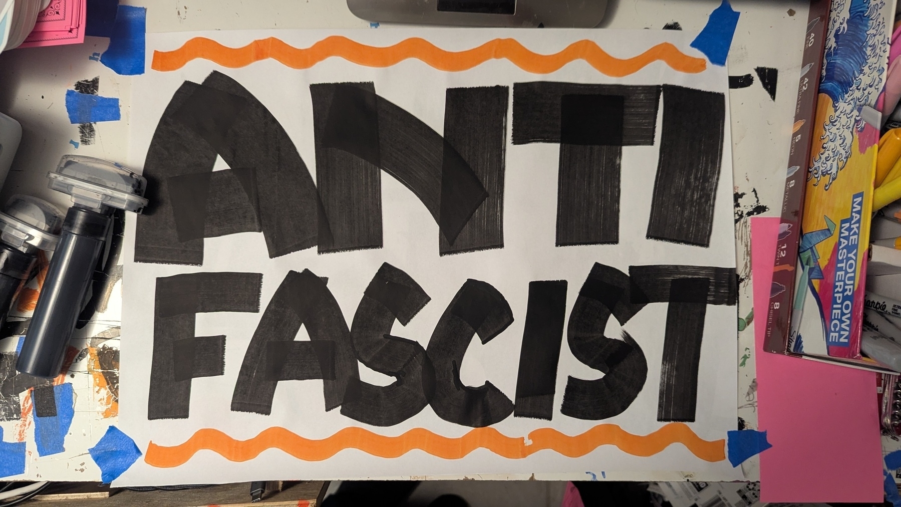 A large handwritten sign reading ANTI FASCIST is displayed on a cluttered table surrounded by art supplies and orange wavy lines framing the words.
