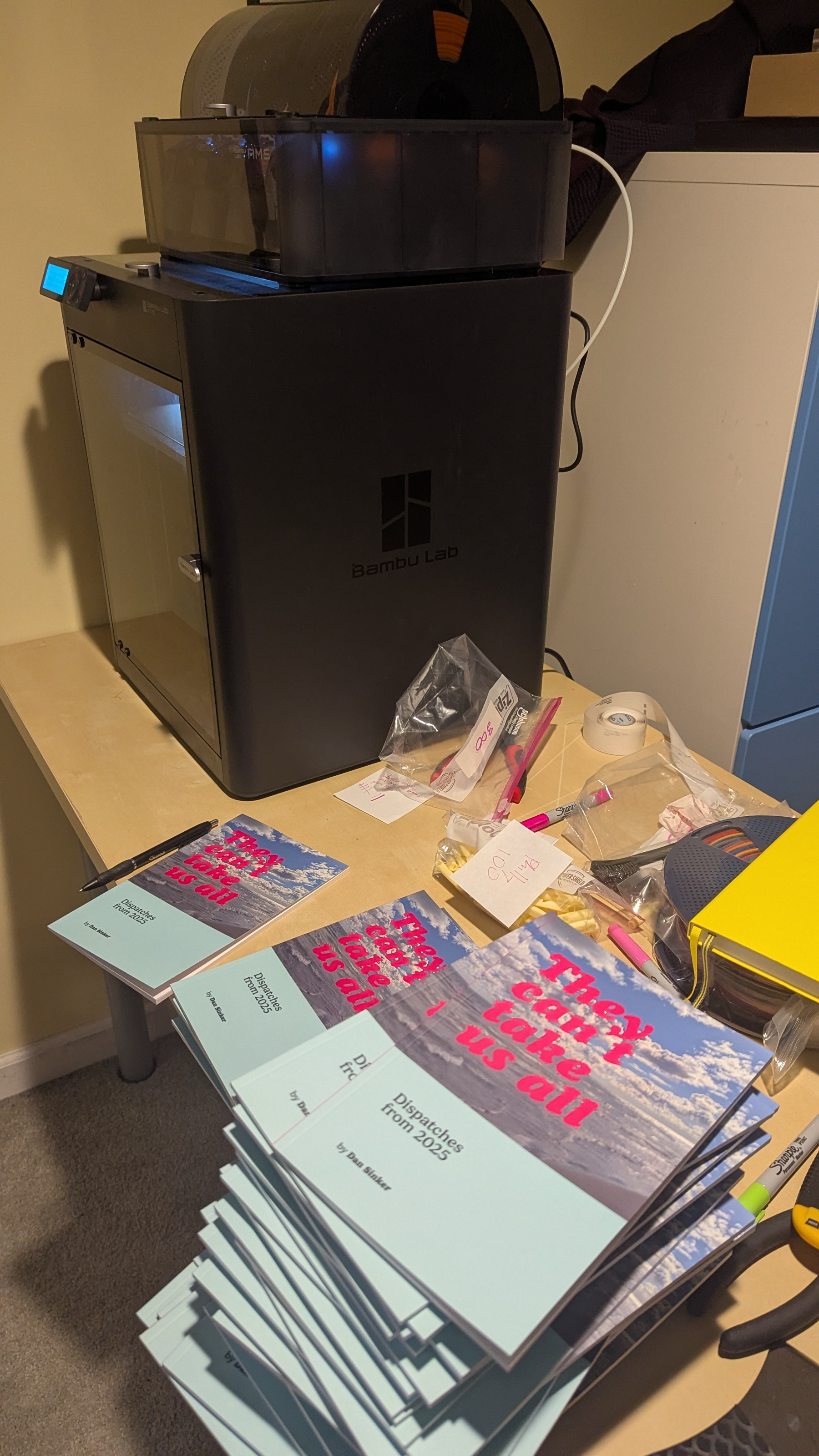 A 3D printer sits on a table with stacks of zines titled "They Can't Take Us All" next to it.