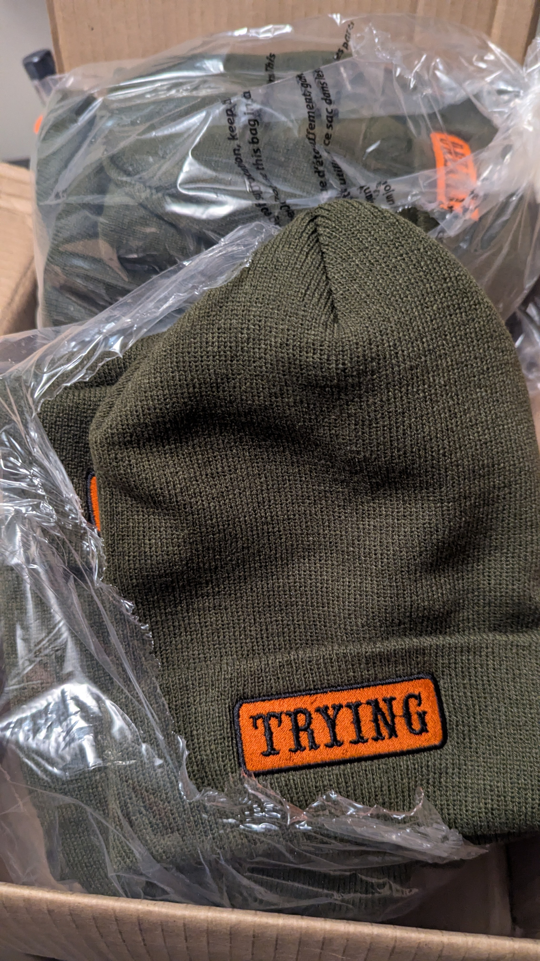 A box contains multiple olive green knit hats with an orange patch labeled TRYING.